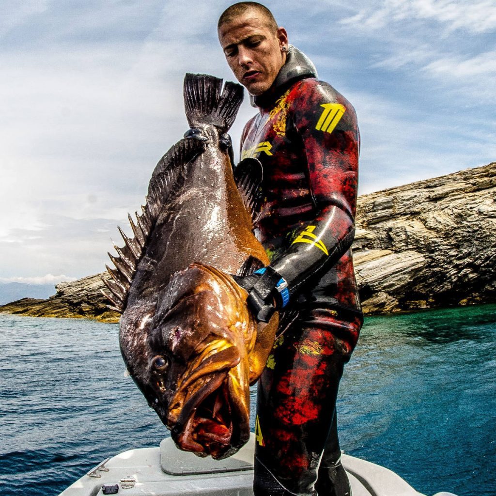 Spearfisher Dimos Adamis on a boat holding a massive Dusky Grouper after a successful dive near rocky shores in Greece.